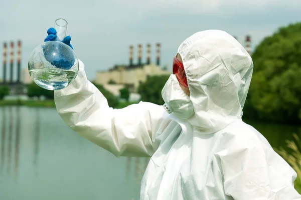 Sampling from open water. A scientist or biologist takes a water sample near an industrial plant. A sample of water in a round chemical flask.