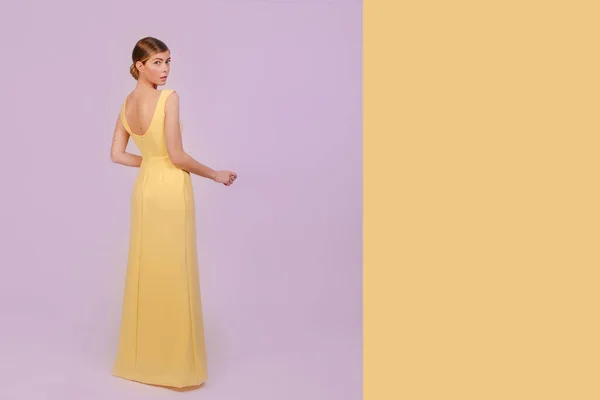 Full shot of beautiful stylish blonde girl in yellow dress looking at the camera presenting your product. Isolated on purple and yellow background. Design copy space.