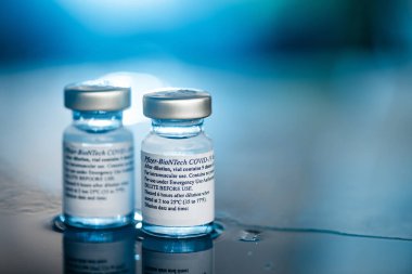 San Sebastian, Gipuzkoa, İspanya; 24 Nisan 2021. İki Covid-19 Pfizer aşı şişesi tıbbi uzmanlara ve masada risk altındaki insanlara enjekte etmek için. Statik askı odağı. Mavi arkaplan ve ışık.