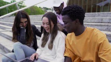 Dört üniversite öğrencisi merdivenlerde oturmuş, okuldaki dizüstü bilgisayara bakarken eğleniyorlar. orta 4k atış