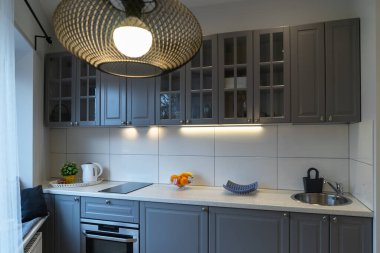 Küçük, rahat, modern bir stüdyo dairede temiz bir mutfak. Pencerenin yanında bir sürü kahverengi dolap, tezgah, lavabo, fayanslı bej duvarlı mutfak aletleri olan yemek pişirme alanı. Ön planda elektrik lambası