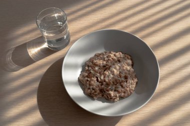 Ahşap masada sağlıklı bir kahvaltı: bir bardak su ve keten tohumlu bir tabak sıcak yulaf lapası, kabak tohumu. Gölgeleri oynamak için harika bir sabah havası. Sağlıklı yemek konsepti. Üst manzara, yakın çekim.