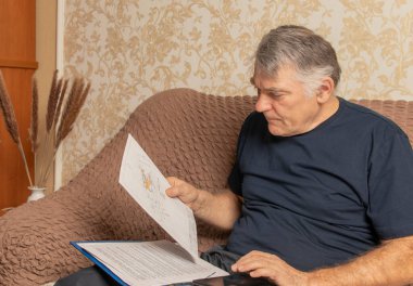 Evdeki kanepede oturan bir adam, belgelerle çalışıyor, posta kağıtlarını inceliyor ve okuyor. Gözlüklü ciddi bir adam evde uzaktan çalışıyor..