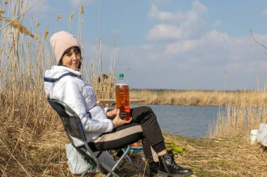 Woman in early spring sits on the river bank on a folding chair, picnic outdoors, selective focus