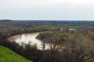 Yeşil bir orman ve yeşil çayır çimenleriyle güzel bir ilkbahar manzarası. Önünde kıvrımlı bir nehrin arkaplanı var. Tarlalar ve tepeler arasında ve mavi, hafif bulutlu gökyüzü..