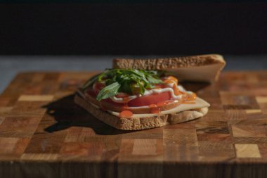 Jambonlu sandviç, roka, peynir ve domates dilimleme tahtasında. Karanlık bir anahtarla. Seçici odak.