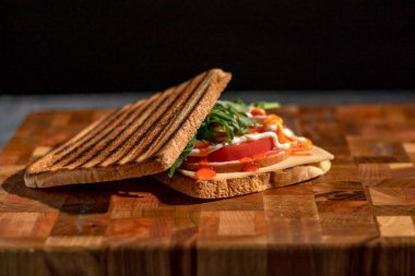 Jambonlu veya balikli lezzetli sandviç, roka, peynir, domates ve mayonez kesme tahtasının üzerinde koyu bir arka plan görünümünde. Seçici odak.