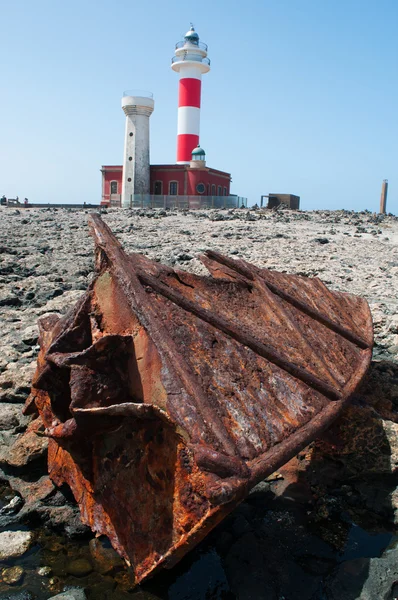 Фуэртевентура, Канарские острова, Испания: чёрные скалы с маяком Faro de Toston (маяк Toston или El Cotillo), действующий маяк в Пунта-де-ла-Баллена (Кит-Пойнт), открытый в 1897 году, видимый сквозь ржавые остатки обломков корабля — стоковое фото