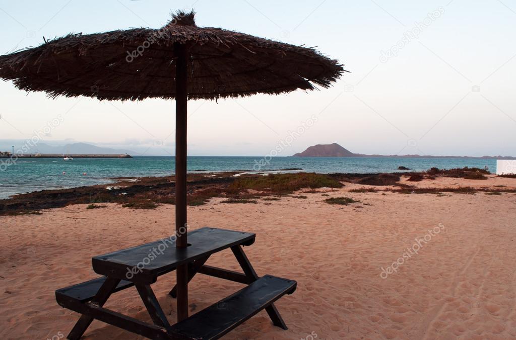 Fuerteventura, Canary Islands, Spain: wooden table, beach umbrella and view of Lobos Island (Islote de Lobos), a small island located 2 kilometers north of the island of Fuerteventura, seen from a beach in Corralejo