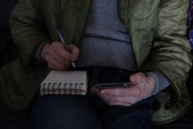 Genç adam akıllı telefon kullanıyor ve not defterine yazı yazıyor. İş planı konsepti. Not defteri tutan iş adamı, akıllı cep telefonu kullanıyor