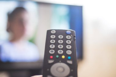 Elinde uzaktan kumanda olan bir TV, ön planda, arka planda bulanık bir televizyon. Akıyor.