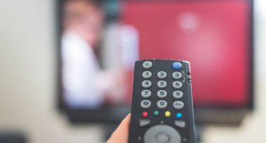 Elinde uzaktan kumanda olan bir TV, ön planda, arka planda bulanık bir televizyon. Akıyor.