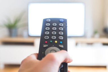 Elinde uzaktan kumanda olan bir TV, ön planda, arka planda bulanık bir televizyon. Akıyor.