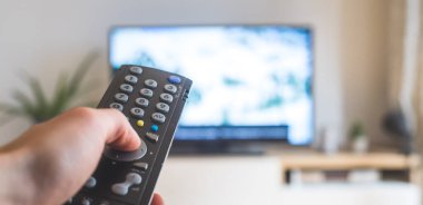 Elinde uzaktan kumanda olan bir TV, ön planda, arka planda bulanık bir televizyon. Akıyor.
