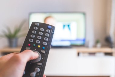 Elinde uzaktan kumanda olan bir TV, ön planda, arka planda bulanık bir televizyon. Akıyor.