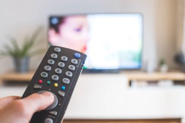 Elinde uzaktan kumanda olan bir TV, ön planda, arka planda bulanık bir televizyon. Akıyor.
