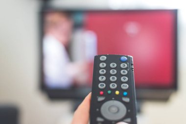Elinde uzaktan kumanda olan bir TV, ön planda, arka planda bulanık bir televizyon. Akıyor.