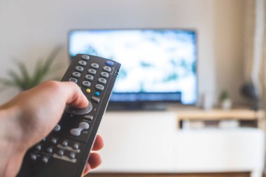 Elinde uzaktan kumanda olan bir TV, ön planda, arka planda bulanık bir televizyon. Akıyor.
