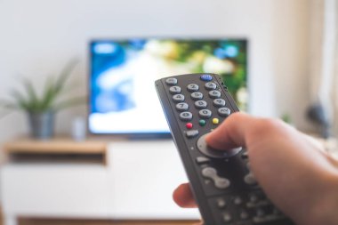 Elinde uzaktan kumanda olan bir TV, ön planda, arka planda bulanık bir televizyon. Akıyor.