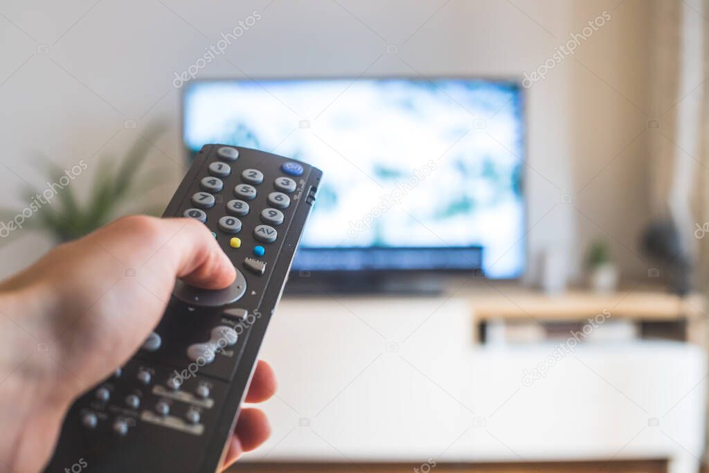 Holding a TV remote control in the hand, foreground, tv in the blurry background. Streaming.