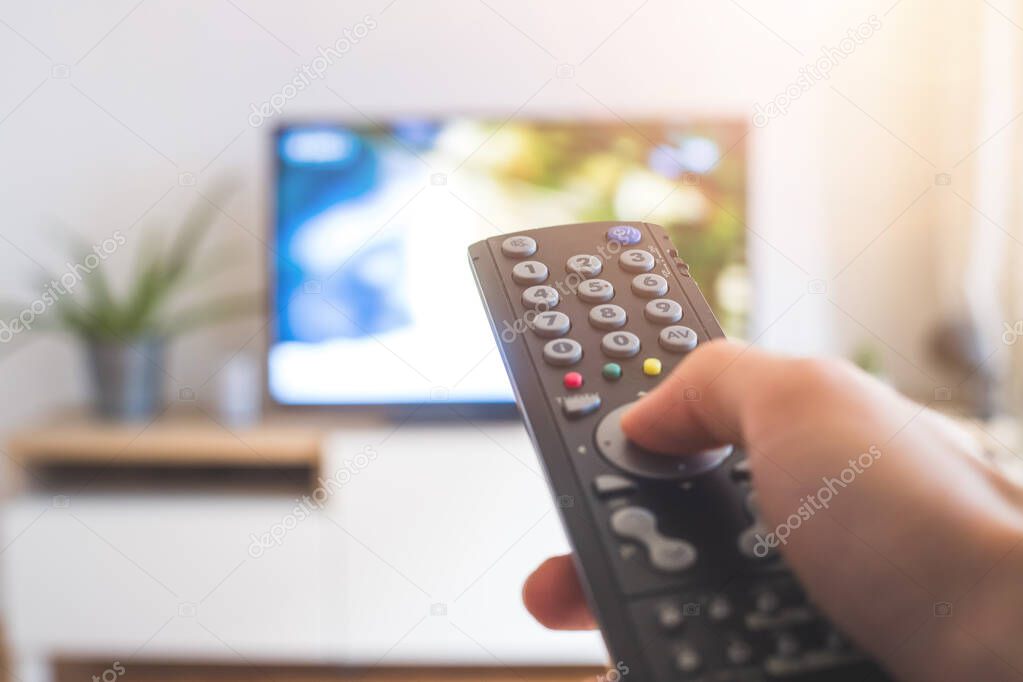 Holding a TV remote control in the hand, foreground, tv in the blurry background. Streaming.