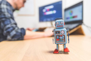 Masaüstünde oyuncak robot, arkada bilgisayarda çalışan adam. Yapay zeka, sohbet robotları veya sosyal robotlar ve algoritmalar için sembol