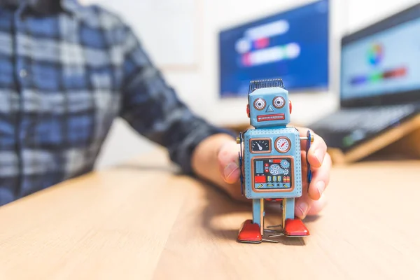 Toy robot on a desktop, man working on the computer in the background. Symbol for artificial intelligence, chatbots or social bots and algorithms