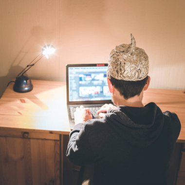 Young man with aluminum cap is sitting in the dark basement in front of a laptop. Conspiracy theory concept clipart