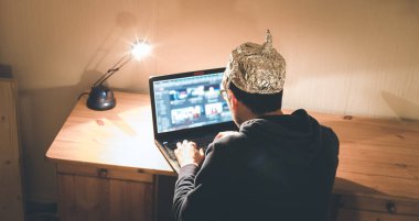 Alüminyum şapkalı genç adam karanlık bodrumda dizüstü bilgisayarın önünde oturuyor. Komplo teorisi kavramı