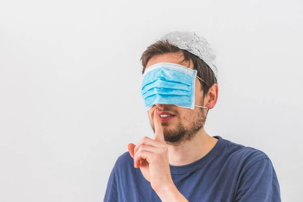 Young angry man with face mask over the eyes and aluminum hat is doing a psst! gesture