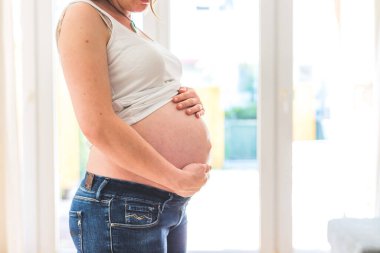 Mutlu Kafkasyalı hamile anne çıplak karnına dokunuyor, kot pantolon, yakın çekim.