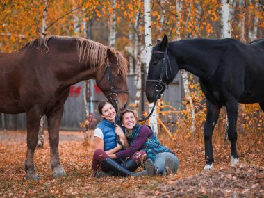 İki kadın binici atların yanındaki çimlere oturur..