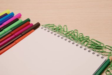 Renkli keçeli kalemler, yeşil ataçlar, kalem ve boş bir defter tahta bir arka planda yatıyor.