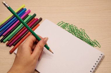 Renkli keçeli kalemler, yeşil ataçlar ve temiz bir defter. Ahşap bir arka planda yatıyor. Bir kadının eli kalemi tutuyor ve deftere yazıyor.