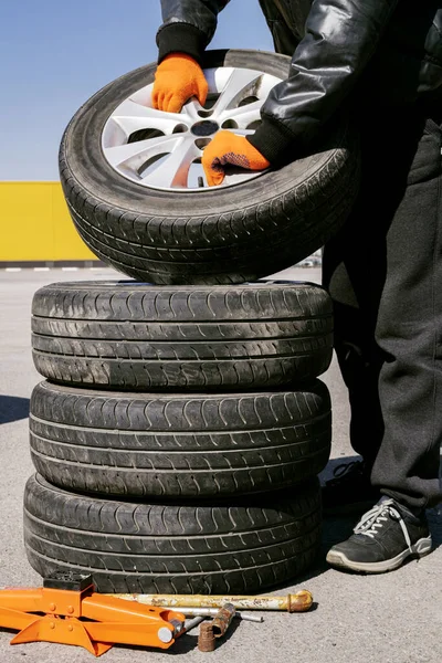 Male auto mechanic holding wheel in orange tire gloves, other three wheels lying on top of each other next to jack and wrenc