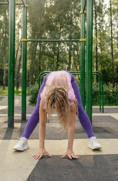 A blonde girl in a sports uniform on the workout site performs stretching of the legs