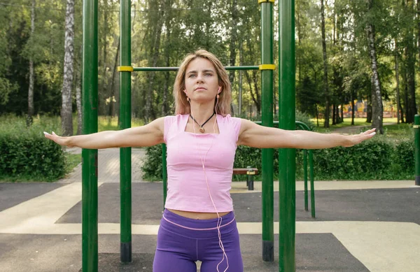 A blonde girl in a sports uniform on a workout playground performs stretching of her arms