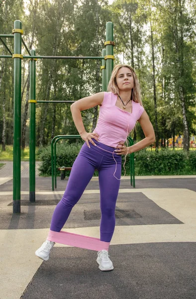 A blonde girl in a sports uniform on a workout site performs an exercise with an elastic band on her legs and buttocks