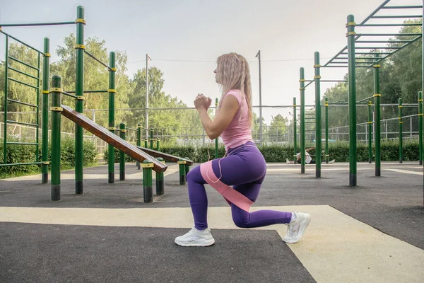 A blonde girl in a sports uniform on a workout site performs an exercise with an elastic band on her legs and buttocks
