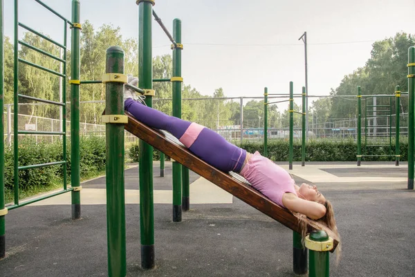 A blonde girl in a sports uniform on a workout playground performs a swing of the abdominal muscles
