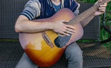 A man, whose face is not visible, plays the guitar on a musical instrument, sitting in nature