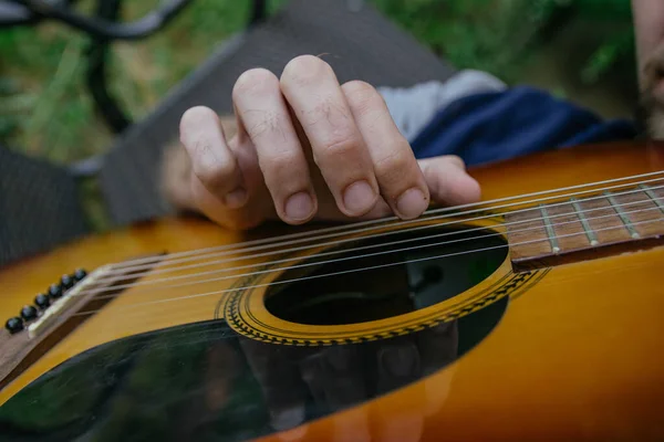 A man, whose face is not visible, plays the guitar on a musical instrument, sitting in nature