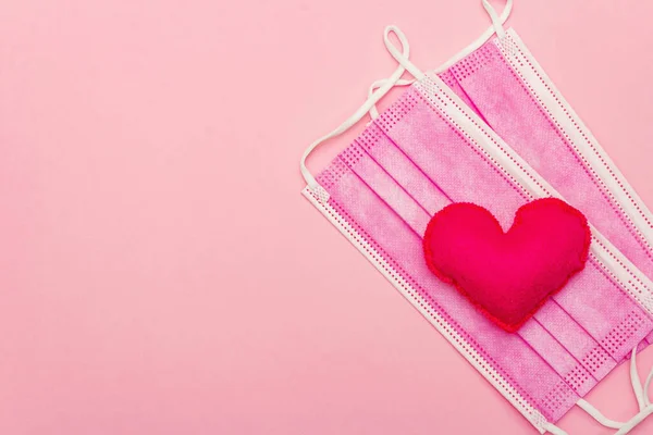 Red heart with medical protection mask on light pink background, top view, copy space. Concept of healthcare, self-defense