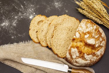 Tahta tezgahta buğday kulaklı taze ekmek. Dilimler, keten peçete, bıçak. Siyah taş arkaplan, üst görünüm