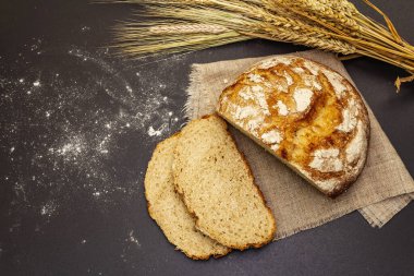 Tahta tezgahta buğday kulaklı taze ekmek. Dilimler ve keten peçete. Siyah taş arkaplan, üst görünüm