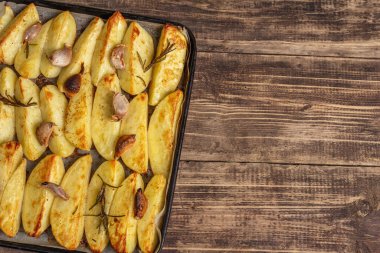 Baked pieces of spiced potato in a baking pan. Fragrant rosemary, garlic, sea salt, oil. Vegan healthy food concept. Vintage wooden boards background, top view