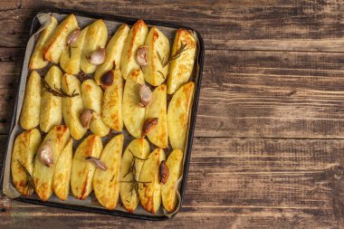 Baked pieces of spiced potato in a baking pan. Fragrant rosemary, garlic, sea salt, oil. Vegan healthy food concept. Vintage wooden boards background, top view