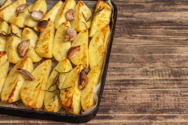 Baked pieces of spiced potato in a baking pan. Fragrant rosemary, garlic, sea salt, oil. Vegan healthy food concept. Vintage wooden boards background, copy space