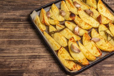 Baked pieces of spiced potato in a baking pan. Fragrant rosemary, garlic, sea salt, oil. Vegan healthy food concept. Vintage wooden boards background, copy space
