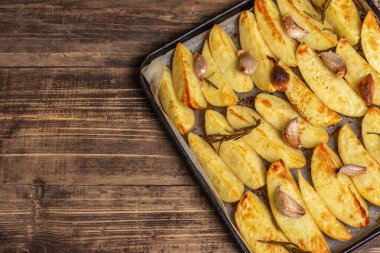 Baked pieces of spiced potato in a baking pan. Fragrant rosemary, garlic, sea salt, oil. Vegan healthy food concept. Vintage wooden boards background, top view